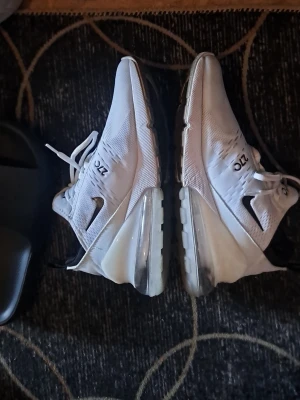 Nike Air Max 270 vita sneakers - Nike Air Max 270 sneakers i vitt med svart swoosh och stora genomskinliga luftkuddar i hälen. Ovandelen är i mesh för bra andningsförmåga och har tydliga Air och 270-detaljer. Skorna har vita snören och svarta detaljer på hälen.