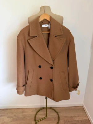 Jacka/kappa - Stilren dubbelknäppt kappa från Ellos i en elegant camel/brun nyans. Klassisk modell med stora slag, svarta knappar, sidfickor och dekorativa detaljer vid ärmsluten.  Storlek 38. Mycket fin och tidlös modell som passar perfekt till både vardag och mer uppklädda outfits.       Ny, aldrig använd