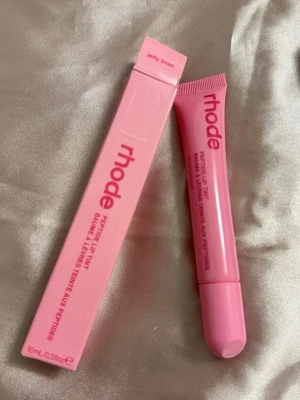 rhode Peptide Lip Tint Jelly Bean - Säljer ett rhode Peptide Lip Tint i färgen Jelly Bean. Läppbalsamet kommer i en rosa tub med matchande förpackning och har en mjuk applikator. Perfekt för dig som vill ha återfuktade läppar med en hint av färg och glossig finish.