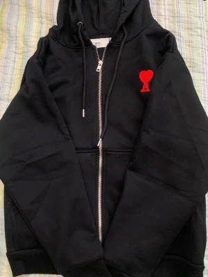 Svart zip hoodie från Ami - Säljer en svart hoodie från Ami med dragkedja framtill och huva med snören. På bröstet finns ett rött broderat hjärta med bokstaven A. Hoodien har fickor på sidorna och ribbade muddar. Perfekt för en chill och stilren look.