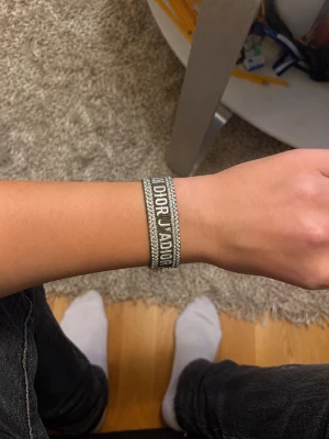 Grönt  J'ADIOR armband med tofsar - Snyggt Grönt armband från Dior med texten 'J'ADIOR' broderat i vitt. Armbandet är gjort av vävt tyg och har dekorativa tofsar i ändarna. Perfekt accessoar för att lyfta din outfit med en exklusiv touch.