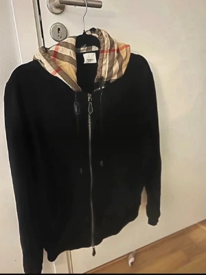 Svart hoodie med mönstrad huva - Säljer en svart hoodie med dragkedja och en huva i klassiskt beige, rött och svart rutmönster. Hoodien har justerbara snören i huvan och ribbade muddar vid ärmar och nederkant. Perfekt för en chill och stilren look.