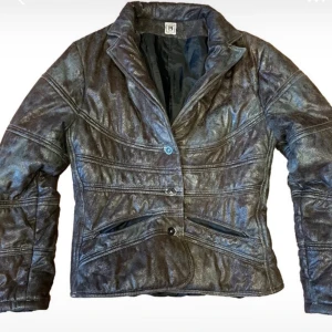 Vintage Brun skinnjacka med knappar - Snygg brun skinnjacka med glansig finish och markerade sömmar. Jackan har två knappar framtill, två snedställda fickor med dragkedja och en klassisk krage. Figurnära passform och coola detaljer som ger en unik look.