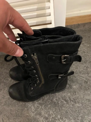 Svarta kängor med spännen och dragkedja - Svarta boots med snörning, dragkedja på sidan och två coola spännen. Grov sula för extra grepp och en edgy look. Perfekta för dig som gillar en tuffare stil. Materialet ser ut att vara syntet eller mockaimitation.