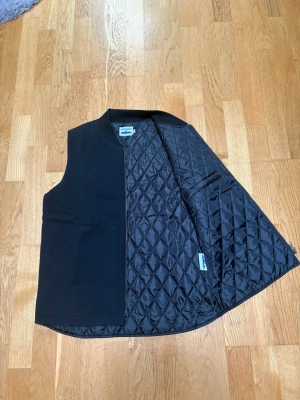 Svart canvas väst - Stilren svart dunväst från Power Goods med quiltat foder och dragkedja. Västen har en enkel, rak passform och ribbad krage. Perfekt att slänga över en hoodie eller tröja för extra värme. Materialet är mjukt och lätt, och designen är klassisk utan onödiga detaljer. Sprillans ny aldrig använd. Nypris 1200kr med frakt och skatt, mitt pris 899kr. 