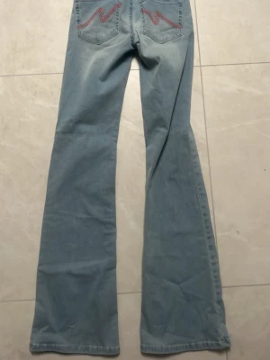Ljusblå bootcut jeans med brodyr - Säljer ett par ljusblå bootcut jeans med rosa broderade detaljer på bakfickorna. Jeansen har klassisk femficksdesign och är tillverkade i denim med lätt stretch. Perfekta för dig som gillar retrovibbar och vill ha ett par riktigt snygga jeans med unik touch.