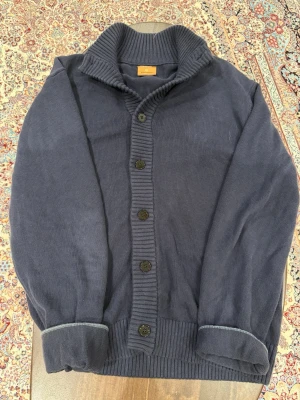 Mörkblå kardigan med krage och knappar - Snygg mörkblå kardigan med bred ribbad krage och knäppning framtill. Koftan har långa ärmar, ribbade muddar och en diskret broderad detalj på ärmen. Perfekt för lager på lager och chill stil.