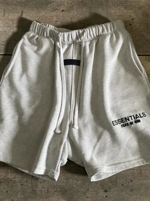 Grå Essentials Fear of God shorts - Snygga ljusgrå shorts från Essentials Fear of God med elastisk midja och snörning. Mjuk bomullskänsla och logga tryckt på vänster ben. Perfekta för sommaren och chill dagar.