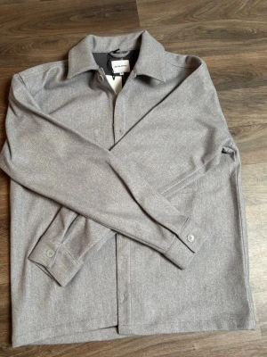 Ljusgrå overshirt från Jack & Jones - Stilren ljusgrå overshirt från Jack & Jones med klassisk krage och knäppning framtill. Tillverkad i ett mjukt tyg som ger en avslappnad look. Perfekt att bära över en t-shirt eller hoodie. Långa ärmar med knappar vid ärmsluten.