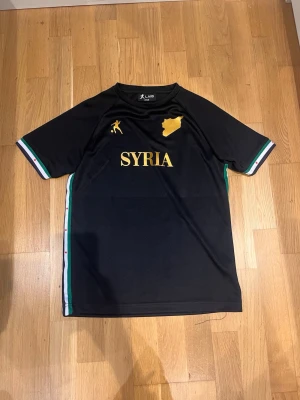Svart Syria fotbollströja LAIB - Säljer en svart fotbollströja med guldiga detaljer från LAIB. Framsidan har texten 'SYRIA' och en siluett av landet i guld, samt en liten fotbollsspelare. Ärmarna har ränder i grönt, vitt och rött. Perfekt för dig som vill stötta Syrien på planen.