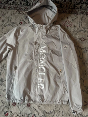Ljusgrå vindjacka från Moncler - Snygg ljusgrå vindjacka från Moncler med stor logga framtill och klassisk Moncler-patch på ärmen. Jackan har huva, dragkedja, tre fickor med dragkedja och justerbar nederkant. Tillverkad i lätt och tåligt material, perfekt för blåsiga dagar.