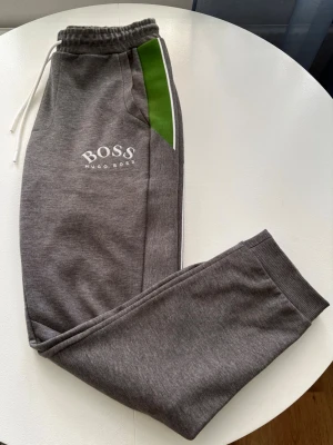 Gråa mjukisbyxor från Hugo Boss - Snygga gråa mjukisbyxor från Hugo Boss med vit logga på vänster ben. Byxorna har vita dragsnören i midjan, gröna och vita detaljer längs sidorna samt ribbade muddar vid bensluten. Perfekta för chill dagar och streetwear-stil. Köpte från Zalando 