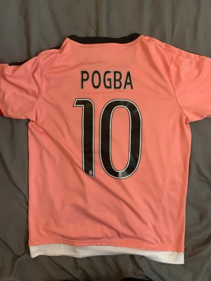 Juventus rosa Pogba matchtröja - Juventus rosa matchtröja med Pogba och nummer 10 på ryggen. Tröjan har svarta detaljer, klubbmärke och Adidas-logga på bröstet samt Jeep som sponsor. Tillverkad i lätt och ventilerande material med svarta ränder på axlarna.