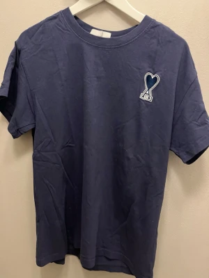 Mörkblå Ami Paris t-shirt - Snygg mörkblå t-shirt med korta ärmar och ett broderat hjärta på bröstet. Tillverkad i mjuk bomull och har en klassisk passform som passar till det mesta. Perfekt för dig som gillar stilrena plagg med en twist.