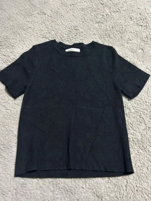 Svart ribbad t-shirt - En stilren svart t-shirt med ribbad struktur och korta ärmar. T-shirten har en klassisk rund halsringning och är tillverkad i ett mjukt material som känns skönt mot huden. Perfekt basplagg för alla tillfällen.