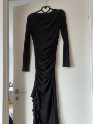 Svart långklänning med rynk och volang - Elegant svart långklänning med långa ärmar och rynkade detaljer längs hela framsidan. Klänningen har en asymmetrisk volangdetalj nedtill som ger extra rörelse och stil. Perfekt för dig som vill sticka ut med en klassisk men unik look.