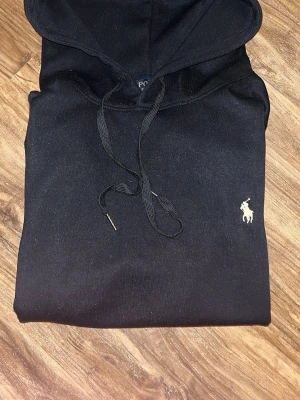 Svart hoodie från Polo Ralph Lauren - Säljer en svart hoodie från Polo Ralph Lauren med klassisk huva och dragsnören. Tröjan har en vit broderad logga på bröstet och en stor magficka framtill. Perfekt för en avslappnad och stilren look.