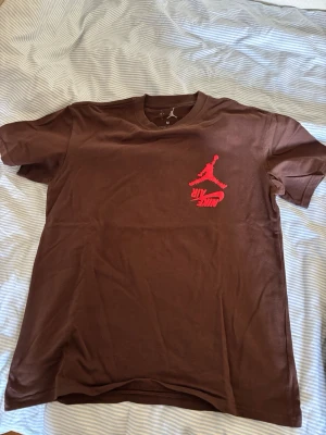 Brun Nike Air Jordan t-shirt - Säljer en brun t-shirt från Nike Air Jordan med röd logga på bröstet. Klassisk rund hals och korta ärmar. Tillverkad i mjuk bomull för skön känsla. Perfekt för dig som gillar streetwear och sportig stil.
