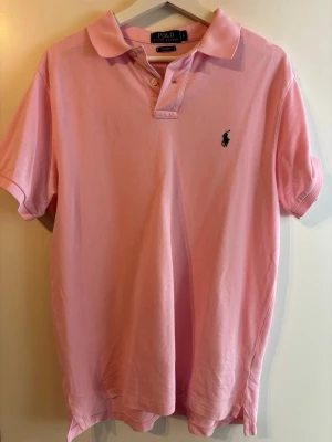 Rosa pikétröja från Ralph Lauren - Säljer en klassisk rosa pikétröja från Ralph Lauren med den ikoniska loggan broderad på bröstet. Tröjan har korta ärmar, krage och knappar vid halsen. Perfekt för en avslappnad men ändå stilren look.