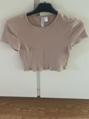 Beige ribbad croppad topp - Säljer en beige croppad topp med ribbad struktur och rund hals. Toppen har korta ärmar och är i ett mjukt, stretchigt material som sitter snyggt på kroppen. Perfekt till jeans eller kjol för en enkel och trendig look.