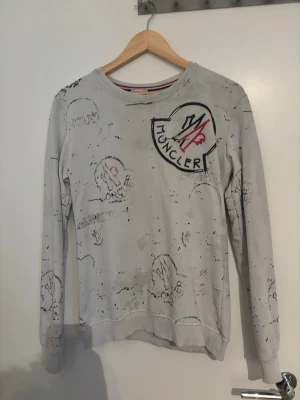Ljusgrå sweatshirt från Moncler - Ljusgrå sweatshirt från Moncler med stort tryckt logomärke på bröstet och abstrakta svarta och röda detaljer över hela tröjan. Rund halsringning och ribbade muddar vid ärmslut och nederkant. Perfekt för en avslappnad och trendig look.