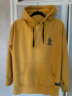 Norrlands hoodie - Säljer en gul hoodie från The Great Norrland med svart tryck av en tecknad figur som sitter på en stubbe. Hoodien har stor känguruficka framtill, ribbade muddar och svart och gult snöre i huvan. På bild 6 ser man bäst hur färgen ser ut i verkligheten. Pruta gärna😝