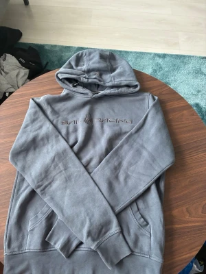 Grå hoodie från Sail Racing - Säljer en grå hoodie från Sail Racing med broderad logga på bröstet. Hoodien har en klassisk huva och känguruficka framtill. Tillverkad i mjukt material som känns skönt mot huden. Perfekt för en avslappnad och stilren look.