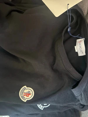 Svart t-shirt från Moncler - Svart t-shirt från Moncler med rund halsringning och klassisk Moncler-logga broderad på bröstet. Enkel och stilren design som passar perfekt till jeans eller shorts.