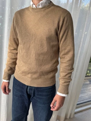 Beige rundhalsad stickad tröja - Klassisk beige stickad tröja med rund halsringning och ribbade muddar vid ärmslut och nederkant. Tröjan har en enkel och stilren design som passar perfekt till jeans eller skjorta under. Materialet ser mjukt och bekvämt ut.