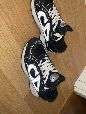 Svarta Dior sneakers med vita detaljer - Snygga svarta Dior sneakers med vita kontrastdetaljer och vita skosnören. Skorna har en chunky sula och meshpartier för extra komfort. Tydlig Dior-logga på plösen och insidan. Perfekta för dig som vill sticka ut med stilrena och exklusiva sneakers. Ordinarie pris på dessa skor är nästan 10 tusen kr så passa på.