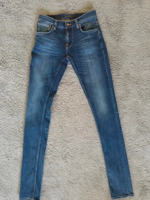 Blå slim fit jeans från Nudie Jeans - Säljer ett par blå slim fit jeans från Nudie Jeans med klassisk femficksmodell och snygga slitningar. Jeansen har normal midja, smala ben och är tillverkade i stretchigt denimtyg för extra komfort. Perfekta för en avslappnad och trendig look.