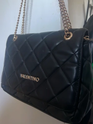 Svart quiltad handväska Valentino - Snygg svart handväska från Valentino med quiltat mönster och guldfärgade kedjehandtag. Väskan har en klassisk rektangulär form och logotypen i guld framtill. Perfekt accessoar för att lyfta din outfit.