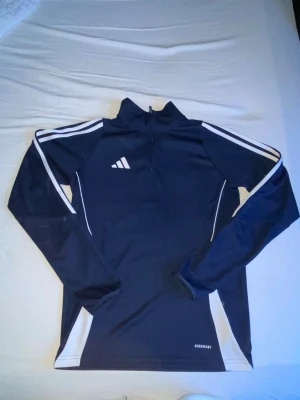 Mörkblå träningströja med dragkedja - Snygg mörkblå långärmad träningströja från Adidas med vita detaljer och dragkedja vid halsen. Tröjan har klassiska vita ränder på ärmarna och är tillverkad i ett stretchigt funktionsmaterial. Perfekt för träning eller uppvärmning.