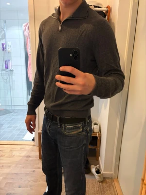 Mörkgrå stickad tröja med halv dragkedja - Mörkgrå stickad tröja med halv dragkedja i halsen och ribbade muddar vid ärmslut och nederkant. Tröjan har en klassisk passform och är perfekt för lager-på-lager. Materialet ser ut att vara mjukt och bekvämt, troligen bomull eller bomullsblandning.