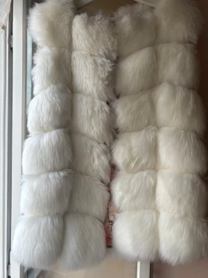 Vit fluffig pälsväst - Supermjuk och lång vit pälsväst med stora, fluffiga paneler och ett snyggt mönstrat innerfoder i rosa toner. Västen har en lyxig känsla och är perfekt för att lyfta vilken outfit som helst. Stängs framtill och har en modern, oversized look.