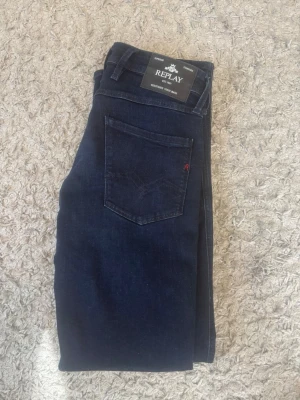 Mörkblå jeans från Replay - Snygga mörkblå jeans från Replay med klassisk femficksdesign och raka ben. Jeansen har en diskret logga på bakfickan och är tillverkade i ett mjukt denimtyg som sitter skönt. Perfekta för dig som gillar stilrena och tidlösa jeans. Aldrig använda.