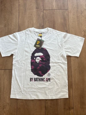 BAPE Camo Ape Head T-shirt i vitt 🐵🟣 (Strl S) - Säljer en skitfet vit Bape-tröja med lila camo-tryck och "By Bathing Ape" på bröstet i storlek S! Använd sparsamt och är i jättefint skick, hängande tags sitter kvar. Inget kvitto finns, men kika in bilderna för detaljer. Skriv ett DM om du undrar över något eller vill ha mått! ✨ 
