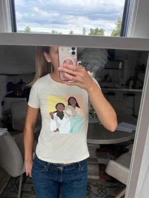 Beige t-shirt med grafiskt tryck - Säljer en beige t-shirt med ett färgglatt grafiskt tryck på framsidan som föreställer tre personer i stiliserad design. T-shirten har rund hals och korta ärmar. Perfekt för dig som gillar unika prints och en avslappnad stil. 
