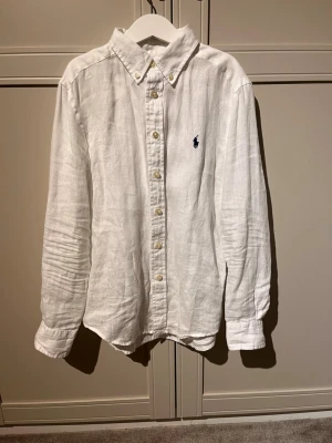 Vit linneskjorta från Ralph Lauren - Klassisk vit skjorta i linne från Ralph Lauren med button-down krage och broderad marinblå logga på bröstet. Skjortan har långa ärmar och pärlemorknappar framtill. Perfekt för en clean och stilren look.