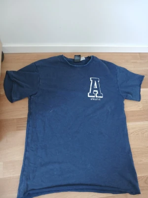 Blå t-shirt med tryck 'A ATHLETIC' - Mörkblå t-shirt med korta ärmar och rund hals. På bröstet finns ett vitt tryck med bokstaven 'A' och texten 'ATHLETIC'. Skön och enkel modell som passar till det mesta. Materialet känns som bomull.
