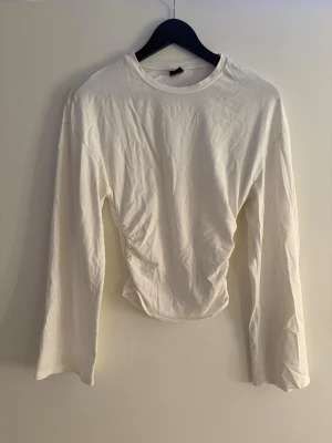 Vit långärmad croppad topp - Säljer en vit långärmad topp med croppad passform och lätt rynkad detalj i midjan. Toppen har rund halsringning och är tillverkad i mjukt bomullsmaterial. Perfekt att styla med höga jeans eller kjol.