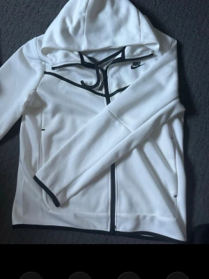 Vit Nike hoodie med svarta detaljer - Säljer en stilren vit hoodie från Nike med svarta detaljer längs dragkedjan, muddar och logga på bröstet. Hoodien har hel dragkedja framtill, två fickor och en skön huva. Perfekt för dig som gillar sportig och clean stil.
