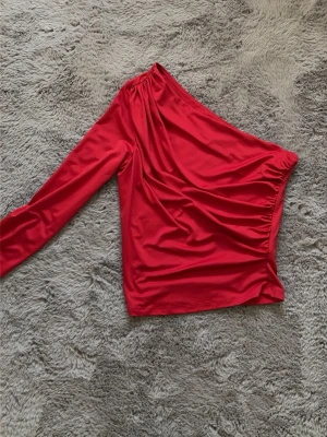 Röd oneshoulder topp från H&M - Snygg röd oneshoulder topp från H&M i stretchigt material. Toppen har lång ärm på ena sidan och är axelbandslös på andra.
