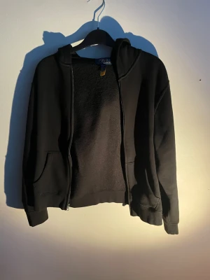 Polo Ralph Lauren hoodie - Svart hoodie med dragkedja från Polo Ralph Lauren. Klassisk design med huva, två fickor framtill och röd broderad logga på bröstet. Tillverkad i mjukt bomullsmaterial som känns skönt mot huden. Perfekt för en avslappnad och stilren look.
