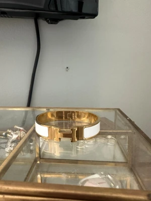 Vitt och guldigt armband med H-detalj - Snyggt armband i vitt och guld med en stor H-detalj framtill. Armbandet har en elegant och stilren design med metallisk finish och vit emalj. Perfekt accessoar för att lyfta din outfit med en lyxig touch.