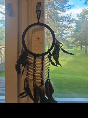 Drömfångare med fjädrar och pärlor - Säljer en snygg drömfångare med svart ring, nät i mitten och dekorationer av bruna och svarta fjädrar samt vita pärlor. Perfekt som dekoration i fönstret eller på väggen. Ger en bohemisk känsla till rummet.