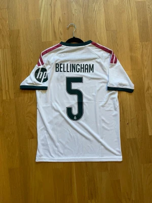Bellingham #5 - M - Säljer en vit Real Madrid matchtröja från Adidas med Bellingham och nummer 5 på ryggen. Tröjan har gröna detaljer vid krage och ärmslut, rosa ränder på axlarna och sponsorloggor på bröst och ärm. Tillverkad i lätt och ventilerande material, perfekt för fotboll.