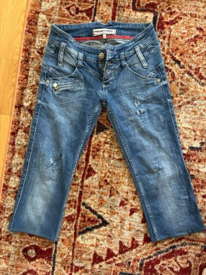 Blå slitna caprijeans Freeman T. Porter - Säljer ett par blå caprijeans från Freeman T. Porter med slitningar och detaljerade fickor fram och bak. Jeansen har låg midja, raka ben och dekorativa sömmar. Materialet är klassisk denim med en cool tvätt och slitna effekter.