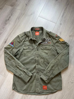 Olivgrön overshirt Superdry patchar - ‼️PRIS KAN ALLTID DISKUTERAS‼️Olivgrön overshirt från Superdry med broderade patchar på bröst och ärm, två bröstfickor med knapp, axelklaffar och knappstängning framtill. Tillverkad i bomull. 