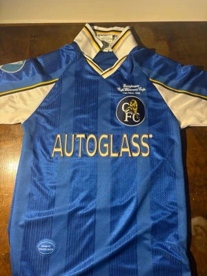 Chelsea retro matchtröja blå - Säljer en blå Chelsea matchtröja med vita och gula detaljer, officiellt klubbmärke och texten 'European Cup Winners Cup The Final 1998' på bröstet. Tröjan har korta ärmar, klassisk krage och är tillverkad i ett glansigt syntetmaterial. Perfekt för samlare eller fans.